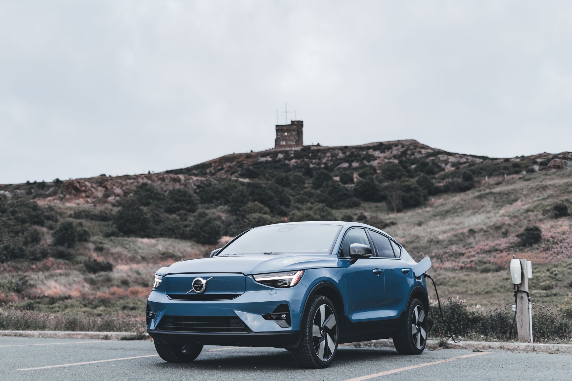 Une voiture électrique Volvo bleue en charge dans un lieu extérieur pittoresque avec une colline en arrière-plan.