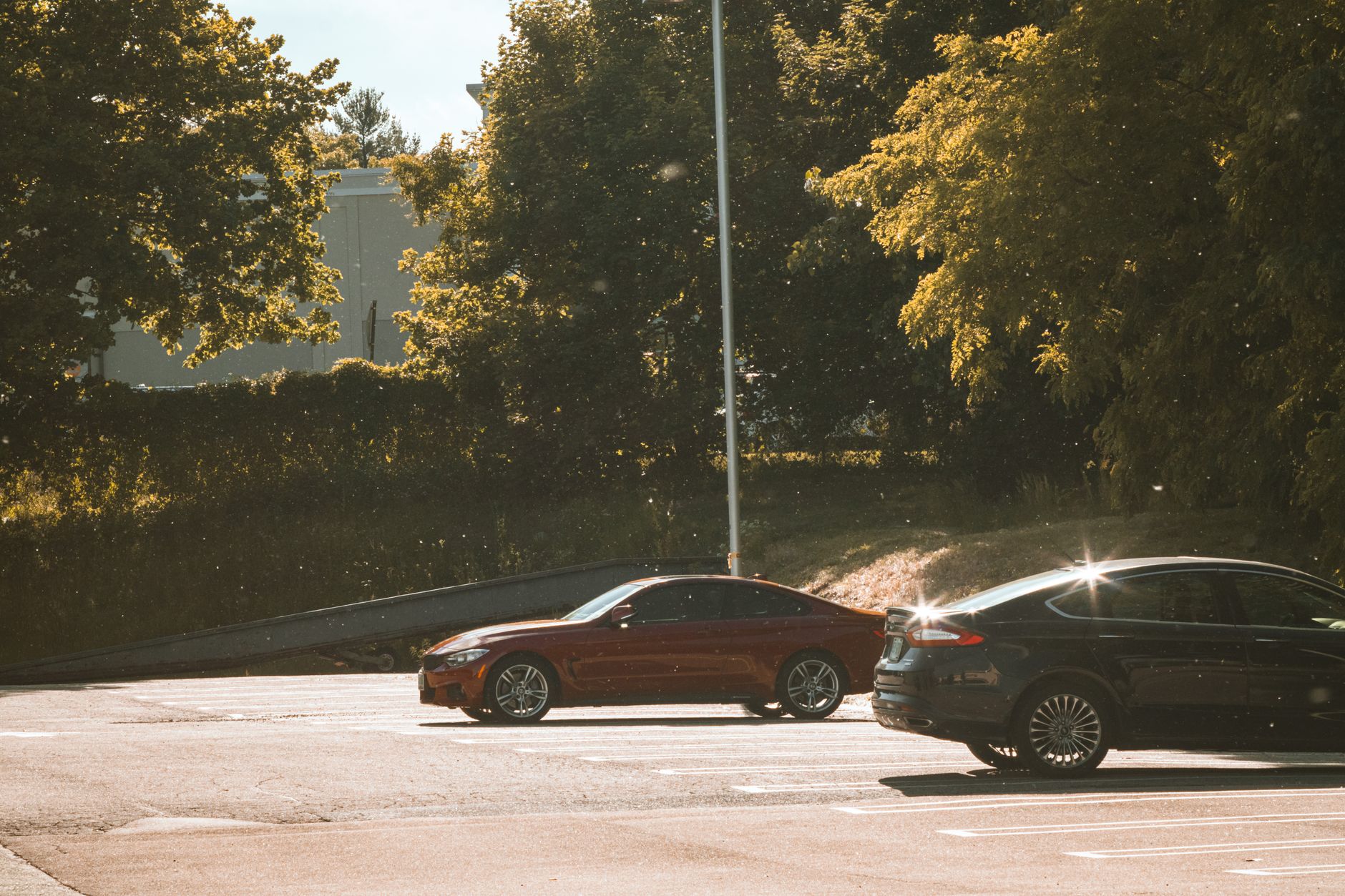 Deux berlines, l’une rouge foncé et l’autre noire, garées sur un parking goudronné ensoleillé devant des arbres et une rampe métallique.