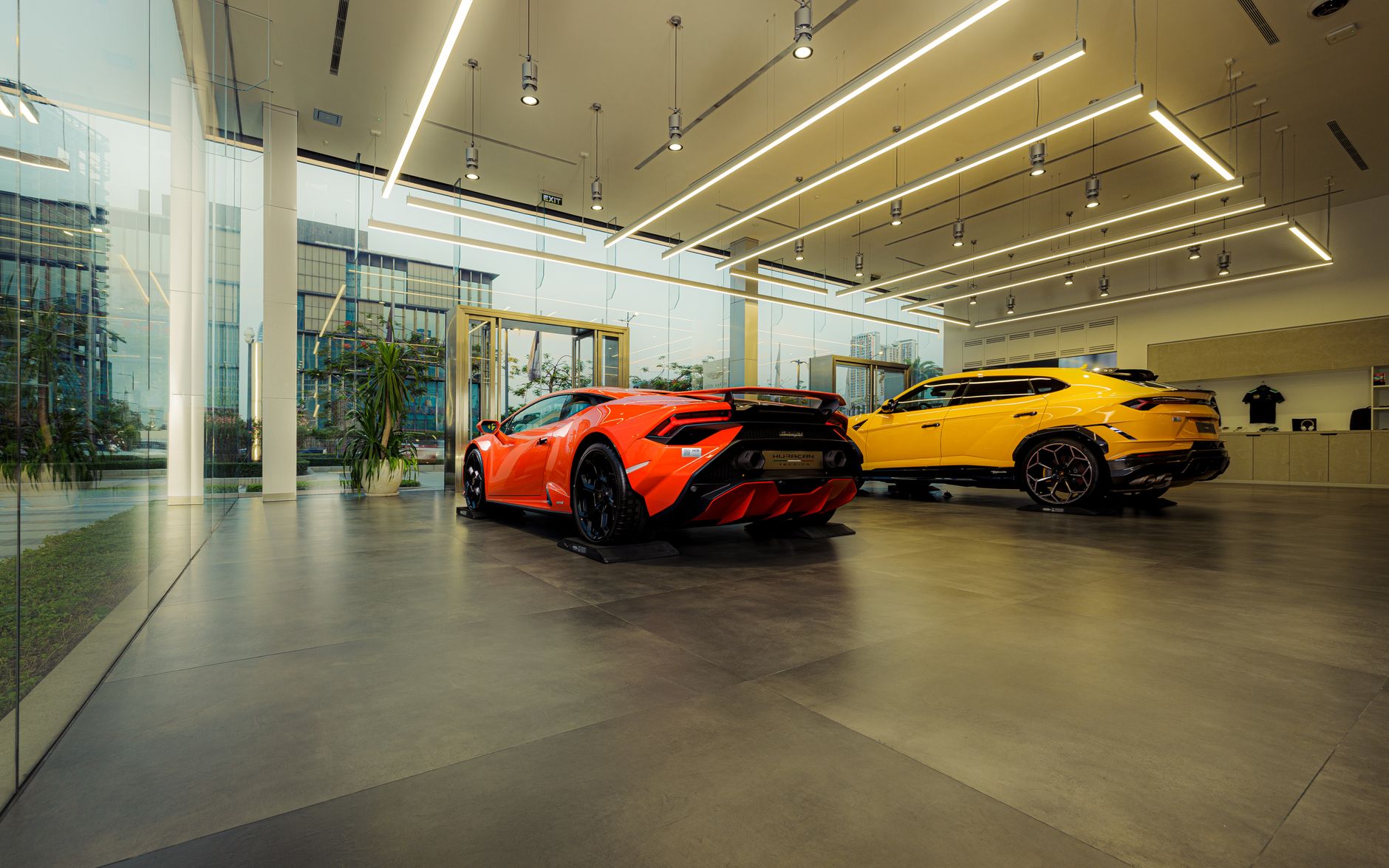 Showroom automobile moderne avec une voiture de sport orange et un SUV jaune, sous un plafond à éclairage linéaire, devant de grandes vitres.