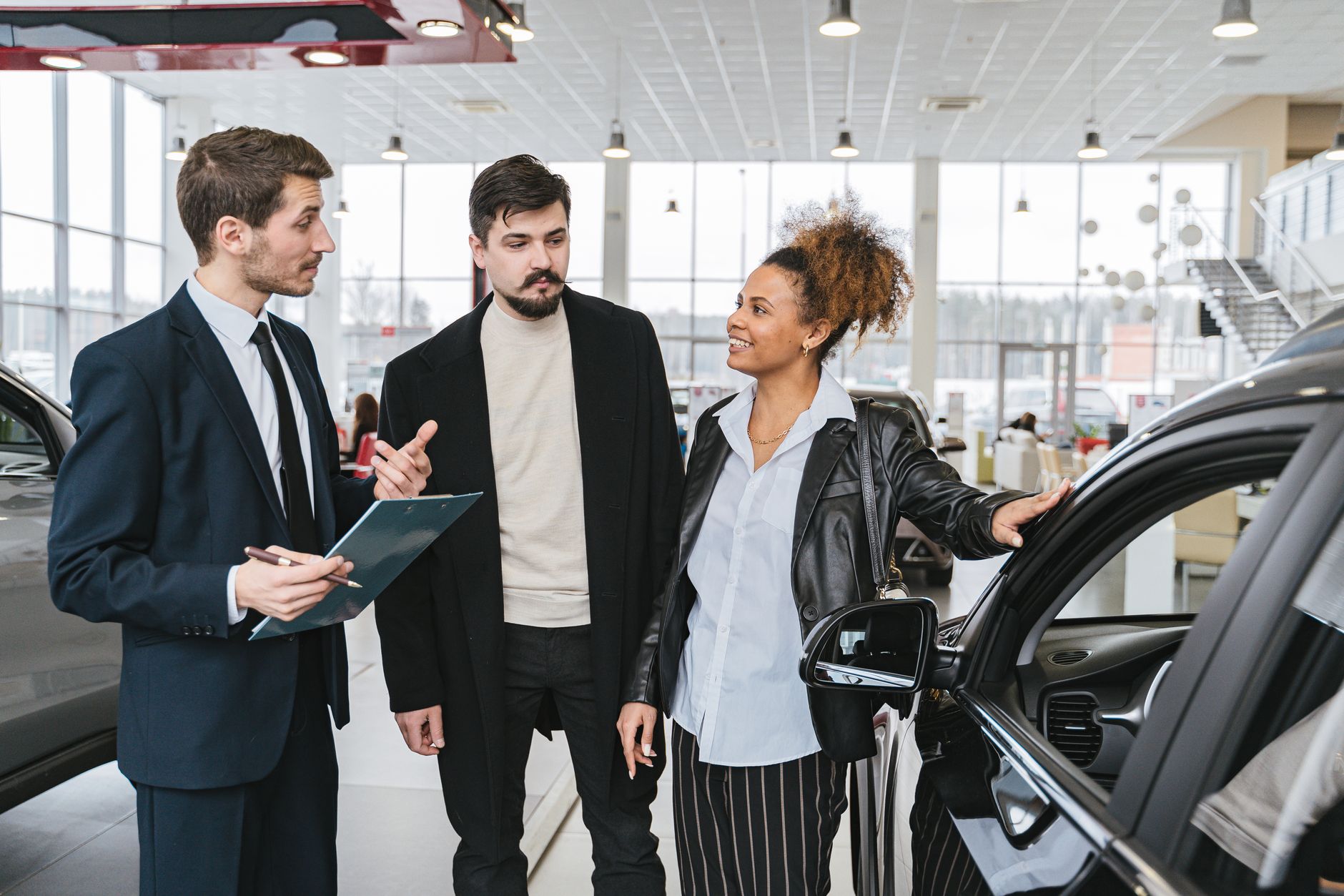 Un vendeur en costume parle à un homme et à une femme près d’une voiture noire dans une concession automobile.
